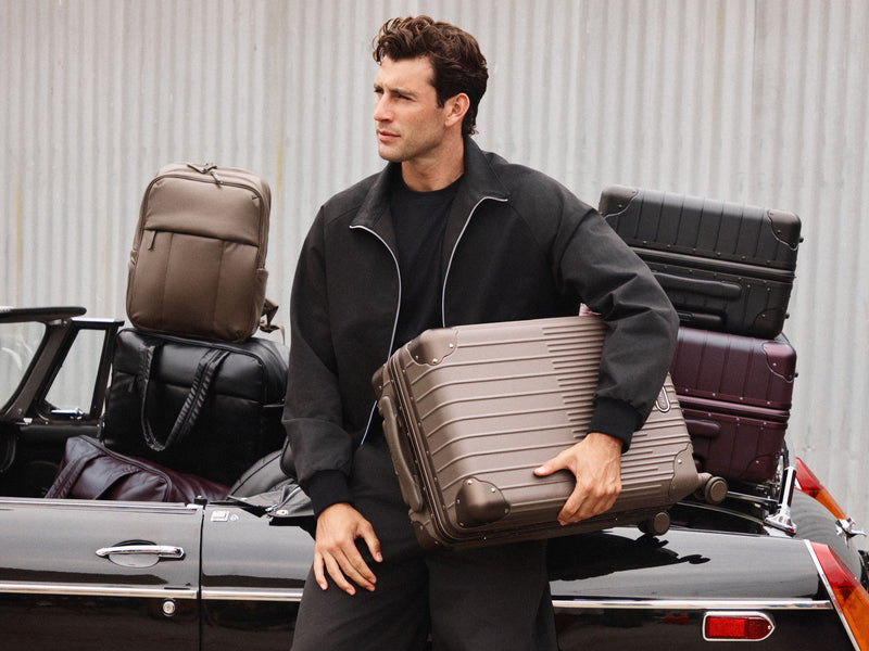 A man in a black jacket stands next to a black convertible car, holding a taupe hard-shell CALPAK Vault luggage. More luggage, including a brown backpack and stacked suitcases in black and burgundy, are piled in the back of the car against a corrugated metal wall.