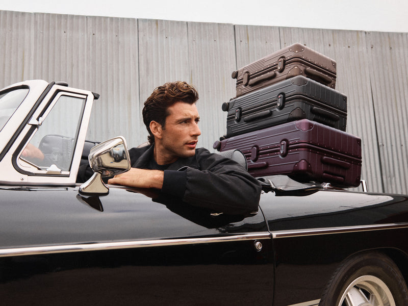 A man leans out of a black convertible car with a stack of four hard-shell CALPAK Vault Collection suitcases piled on the roof against a corrugated metal wall.