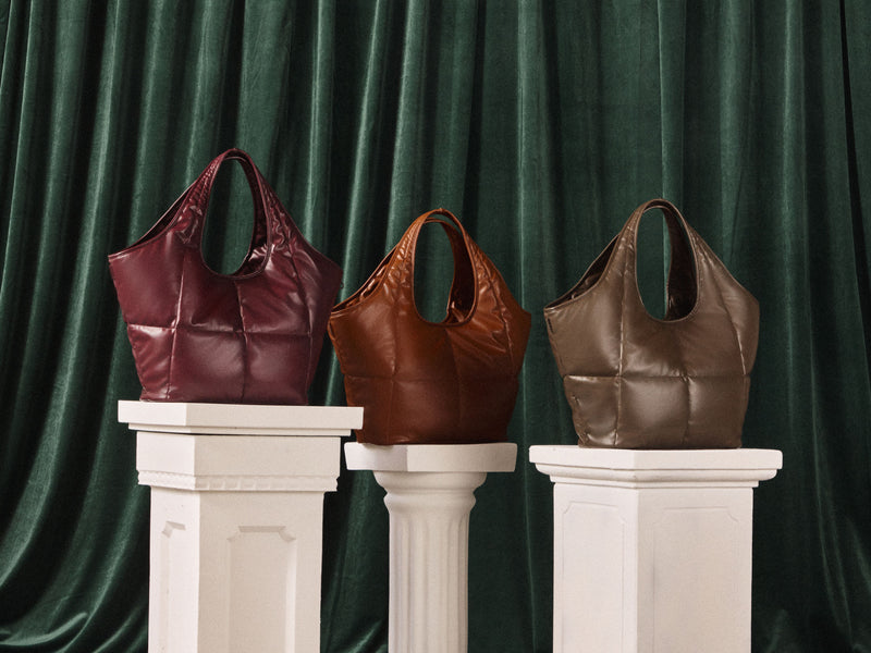 Three large slouchy Luka Everything Laptop Tote bags in burgundy, brown, and taupe vegan leather, displayed on white pedestals against a dark green velvet curtain background.