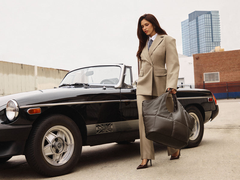A woman in a beige tailored suit stands beside a black convertible car, holding a large dark gray vegan leather Luka Everything Laptop Tote Bag in an outdoor urban setting.