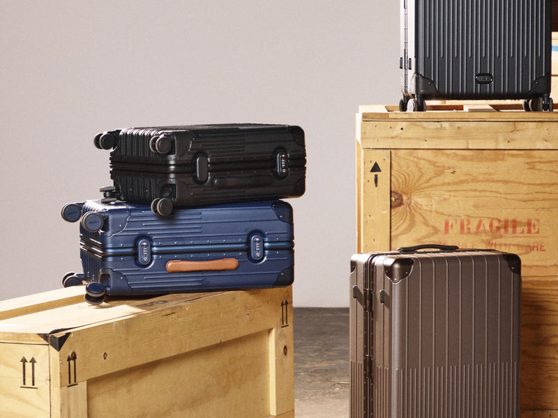 Four hard-shell CALPAK Vault Collection carry-on suitcases in black, blue, taupe, and dark gray, styled on and next to wooden crates against a neutral studio background.