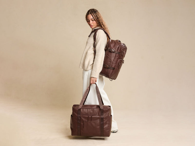 Woman carrying a brown Terra 35L Water Resistant Zippered Tote bag and Terra 26L Laptop Duffel backpack on a beige background
