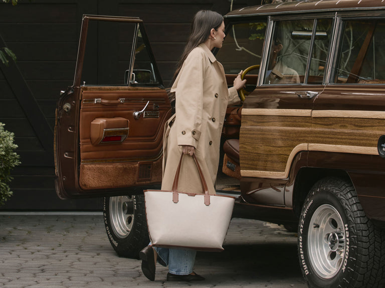Woman in a trench coat getting into a vintage wooden SUV with a white Colette Laptop Tote bag.