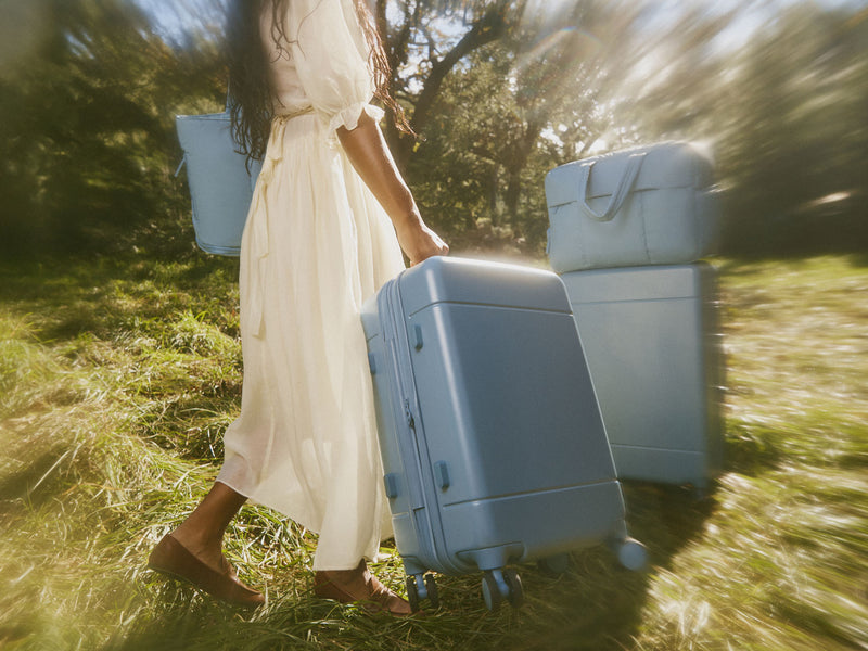 Woman in a white dress walking through a grassy field with two blue CALPAK Hue Collection suitcases.