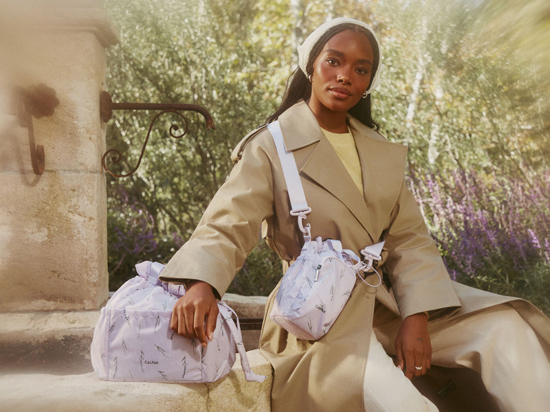 Woman in a beige coat holding a floral patterned CALPAK insulated lunch bag and water bottle holder in an outdoor setting with greenery.