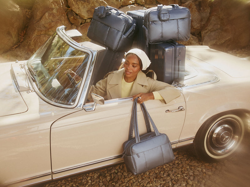 Woman sitting in a vintage car with CALPAK metallic indigo luggage and bags surrounded by rocky terrain