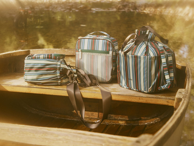 Striped insulated lunch bag, water bottle holder and packing cube on a wooden boat with a blurred natural background