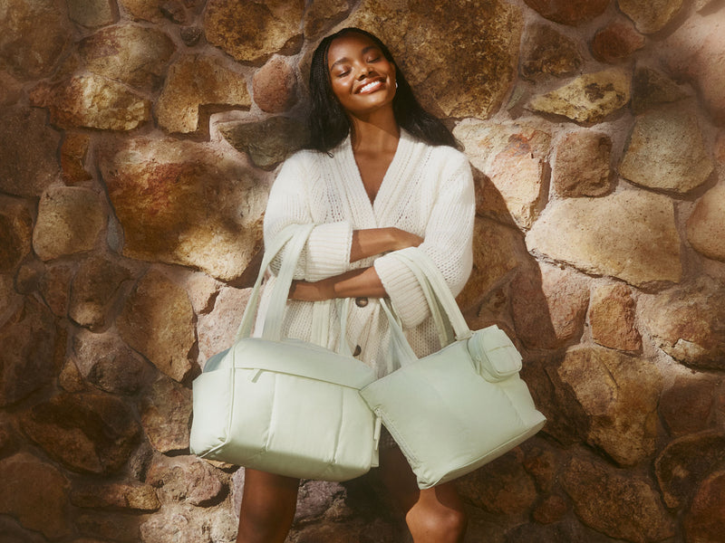 Woman holding two light green CALPAK Luka Collection handbags against a stone wall