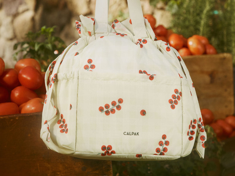 White CALPAK lunch bag with red tomato pattern in front of a fruit stand