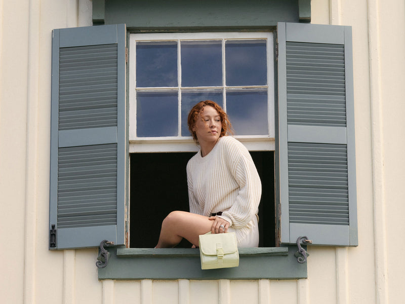 Woman sitting in an open window with a blue shutter, holding the CALPAK Terra Hanging Toiletry bag in light green Celadon