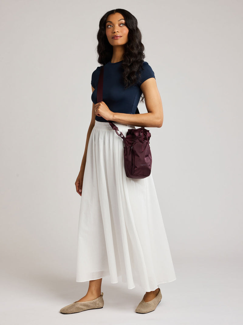 A woman modeling a navy blue short-sleeve top, a long white pleated skirt, and beige flats, carrying a dark burgundy CALPAK Water Bottle Holder with top handles, drawstring top, front zippered pocket, and a detachable shoulder strap