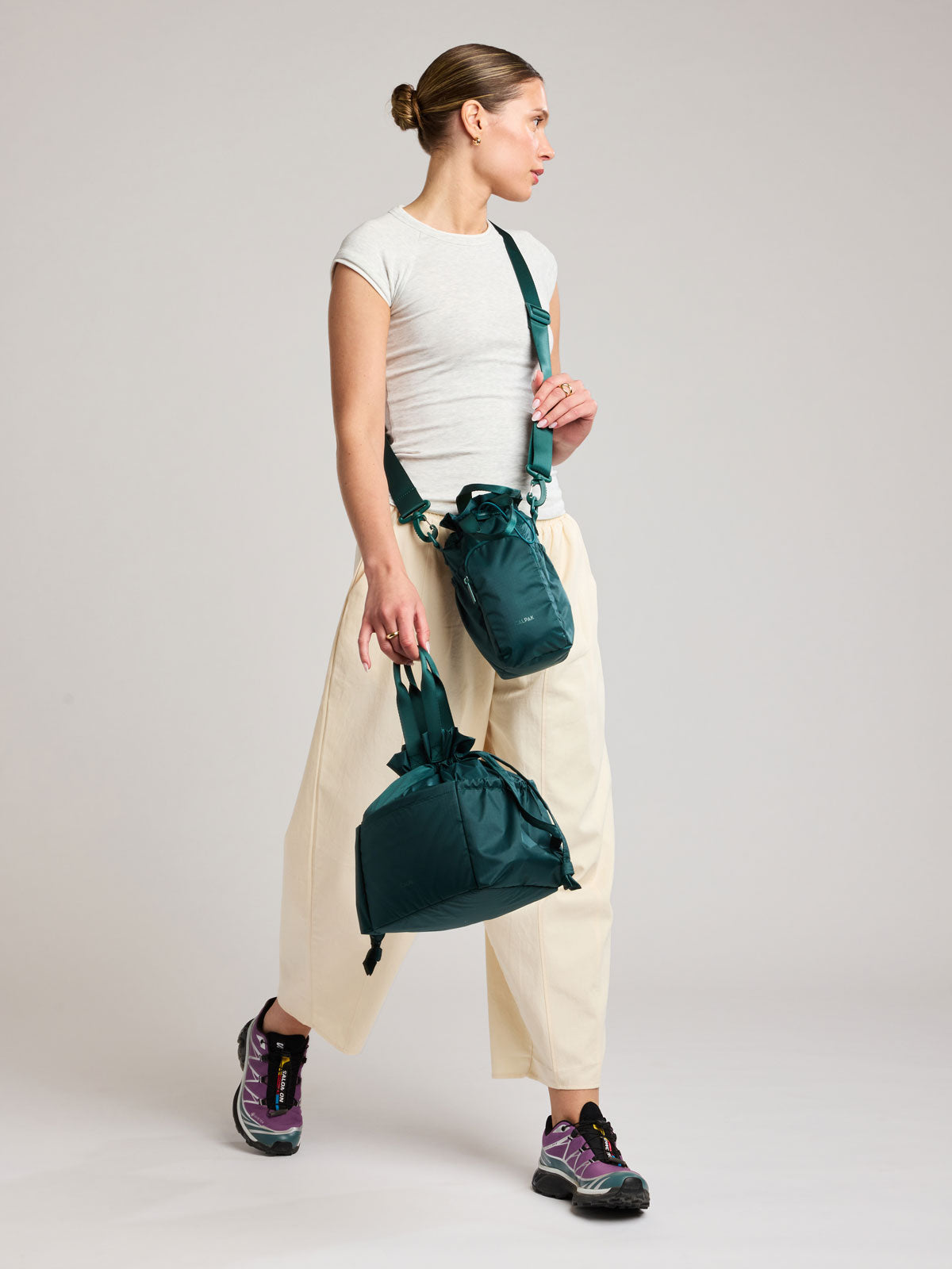 A woman in a white t-shirt and cream wide-leg pants models a dark green teal Kale CALPAK Water Bottle Holder with top handles, drawstring top, front zippered pocket, and a detachable shoulder strap; AWH2101-KALE