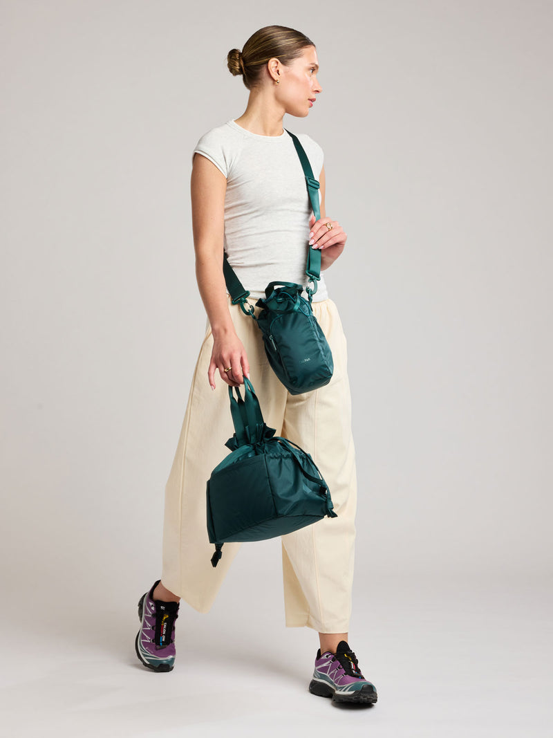 A woman in a white t-shirt and cream wide-leg pants models a dark green teal Kale CALPAK Water Bottle Holder with top handles, drawstring top, front zippered pocket, and a detachable shoulder strap