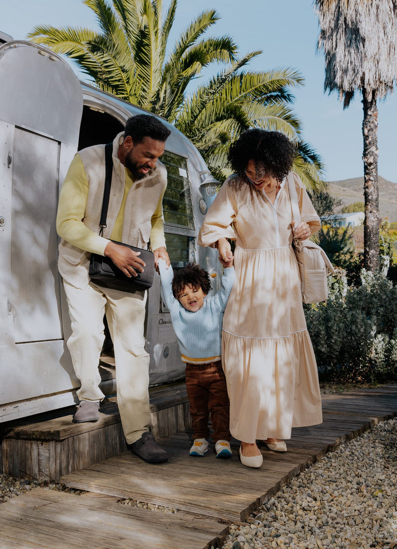 A stylish family steps out of an Airstream trailer into the sunshine, with the father carrying a black CALPAK diaper bag crossbody. The mother holds their toddler’s hand as they walk together on a wooden path surrounded by greenery carrying the CALPAK convertible mini backpack in oatmeal.