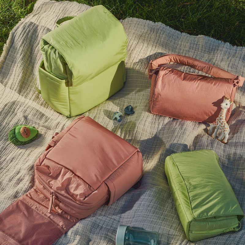 A soft green and blush pink CALPAK diaper bags displayed on a neutral picnic blanket in the grass. The set includes a backpack, crossbody bag, changing pad, and pouches, alongside baby essentials like a pacifier and teething toy.