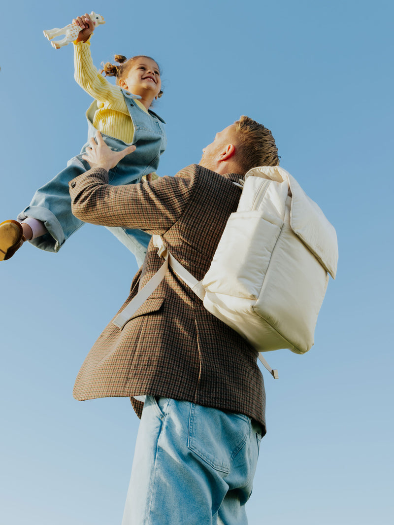A father lifts his joyful daughter into the air against a bright blue sky. He carries a sleek, cream-colored CALPAK diaper backpack, blending style and function for on-the-go parenting.