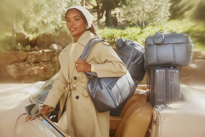 Woman in a trench coat holding a CALPAK Luka Duffel in Indigo blue next to a car with suitcases on a scenic road.