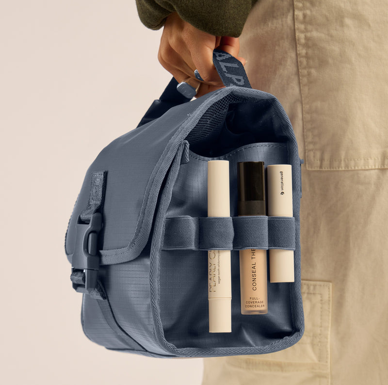 Person holding a blue CALPAK Terra Hanging Toiletry Bag with skincare products inside, wearing beige pants and a green cap.