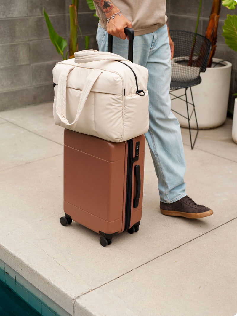 CALPAK Hue Carry On Luggage in brown Hazel being rolled by model with a white Oatmeal Luka Duffel on top of luggage