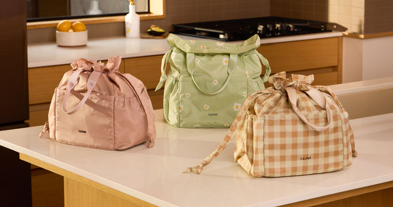 CALPAK Insulated Lunch bags on kitchen counter. Order from left to right is pink Mauve, light green with daisy print Daisy, and tan and brown checkered Gingham.