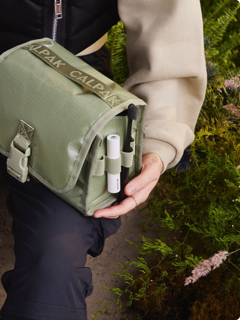 Model holding green CALPAK Terra Hanging Toiletry Bag, showing side view with lip balm and toothbrush