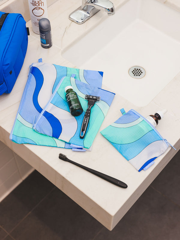 From CALPAK's Compakt collection, these 'Groovy Blue' zippered nylon pouches are displayed on a bathroom sink, offering stylish organization for your essentials